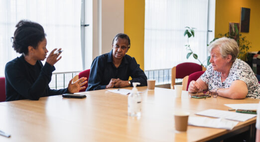 A group of three people sitting at a table. They are working and there are papers, coffee cups and mobile phones in front of them.