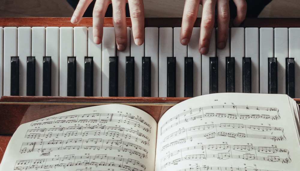 Hands on a piano keyboard behind a musical notation book.