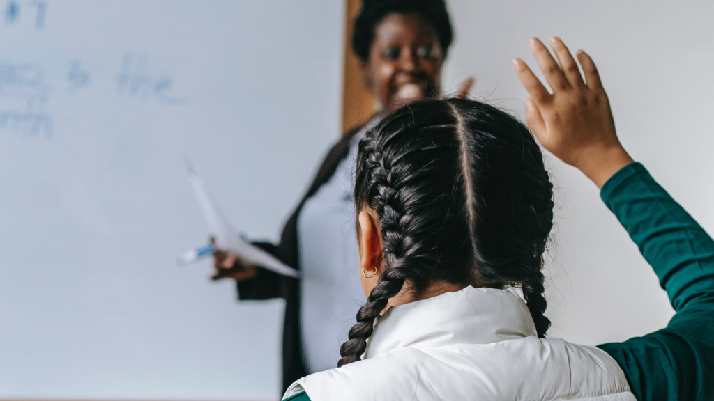 Teacher standing by a white board, and pupil with hand in the air.